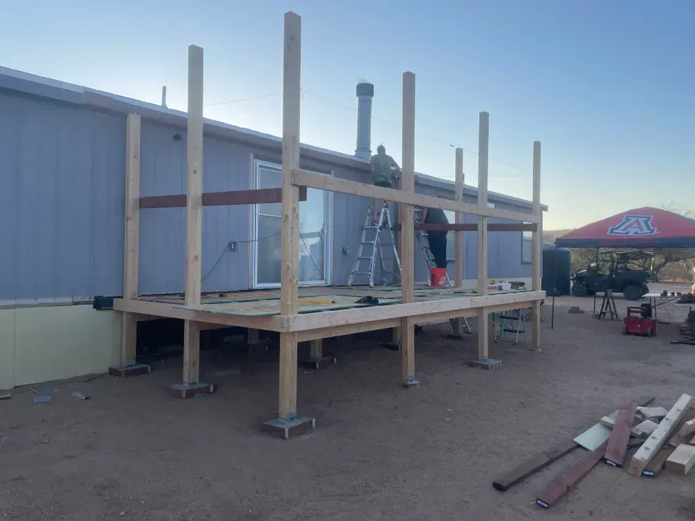 Deck construction in progress; wooden frame built against a building, two people working.