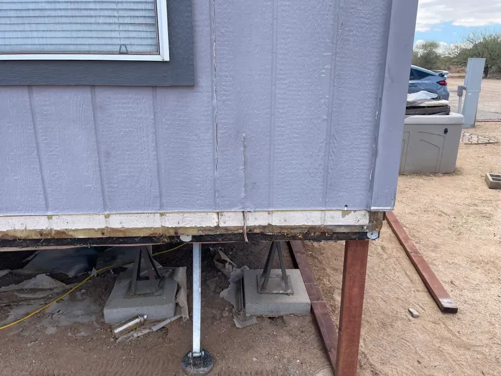 Gray mobile home exterior, showing siding, window, and exposed foundation with support jacks.