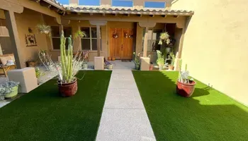 A house entrance with a pathway and potted cacti. Green turf surrounds the path; wooden door.