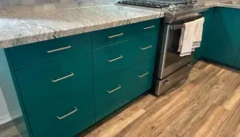Teal kitchen cabinets with silver hardware and a light granite countertop. A stainless steel oven sits to the right.