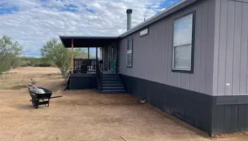 Gray house with a porch and steps, dark gray skirting. Wheelbarrow in yard.
