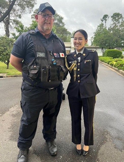 Man in tactical vest stands beside person in military uniform on a paved road.