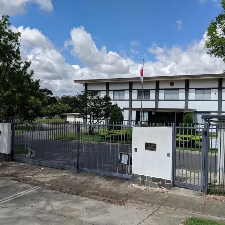 Japanese embassy building with flag flying, open gate, gray exterior, under blue sky.