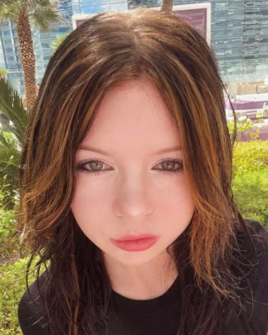 Woman with brown hair, streaks of blonde, looking directly at the camera with a neutral expression. Outdoors, sunny day.