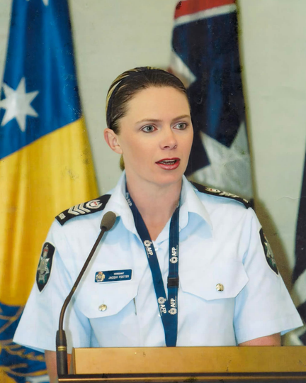 Woman in uniform speaking at a podium, flags in the background.
