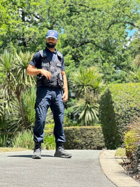 Person in uniform stands on a path, outdoors with greenery. He wears a navy uniform and tactical vest.
