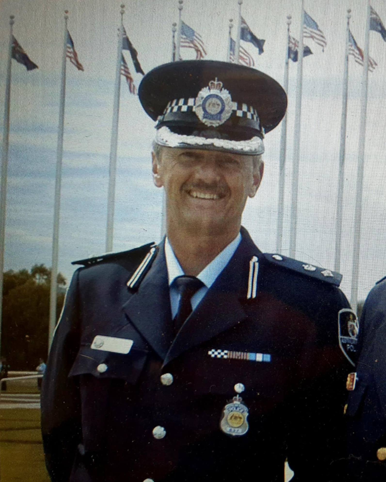 A uniformed police officer with a wide smile, wearing a hat, standing in front of flags.