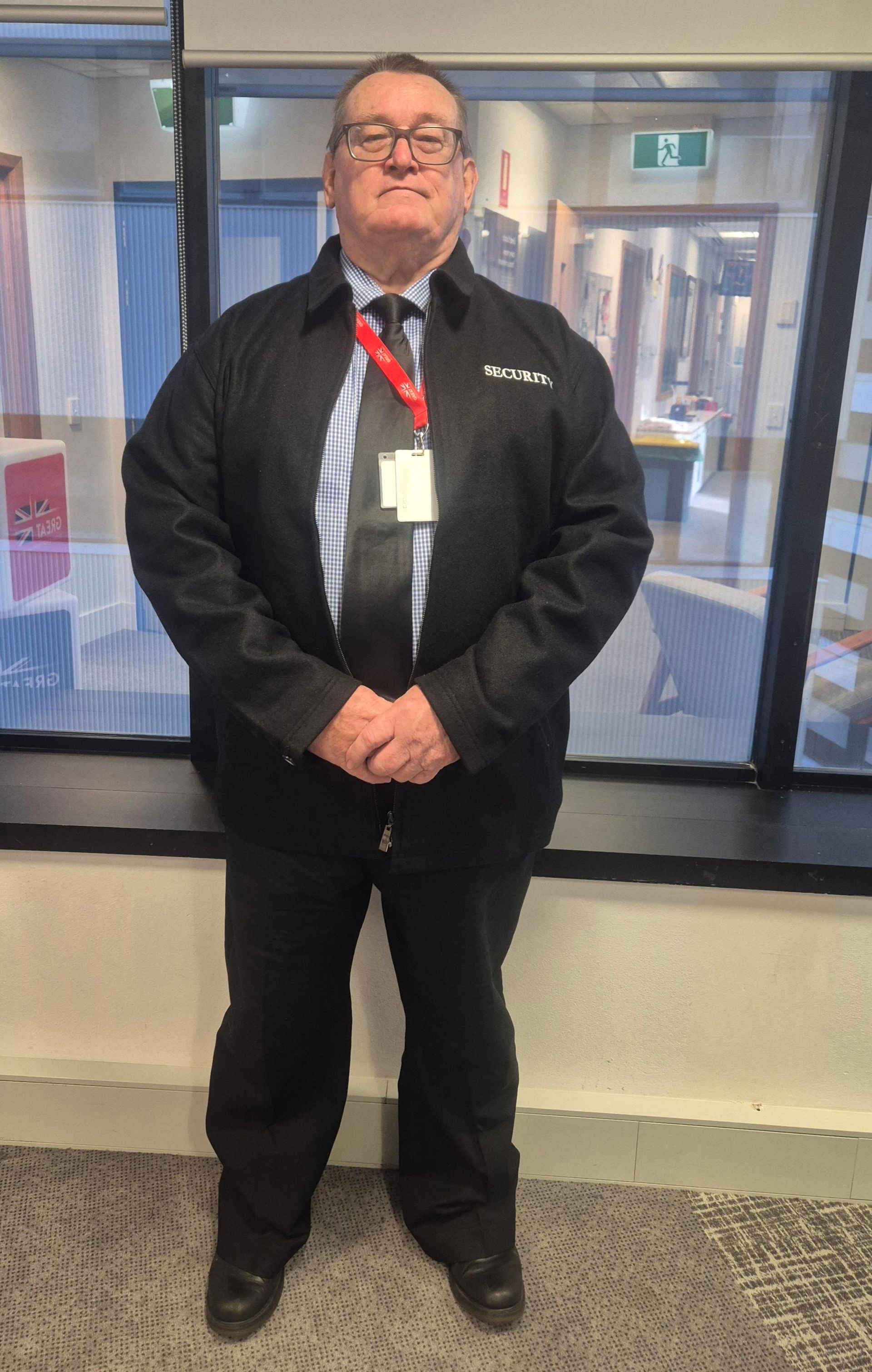Man in security uniform, standing indoors with hands clasped. Black jacket, pants, and tie. Window in background.