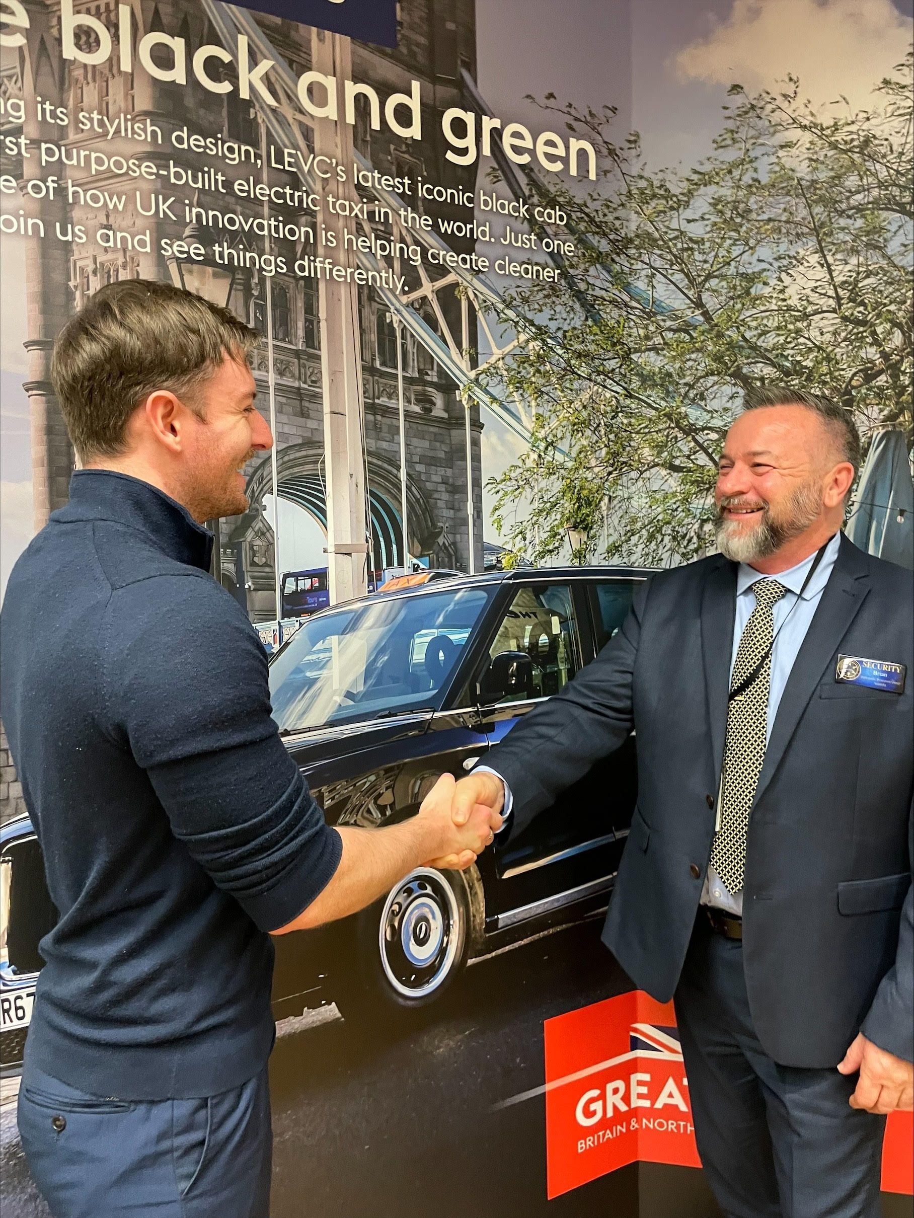 Two men shaking hands in front of a black car graphic; smiles, business attire, indoors.