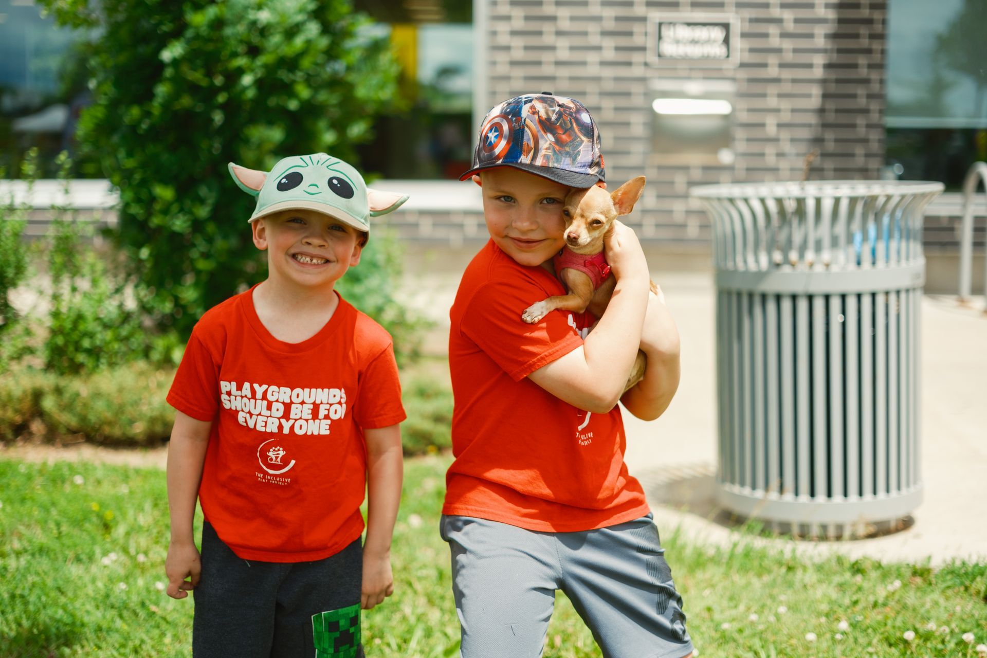 Two young boys are standing next to each other in a park holding a small dog.