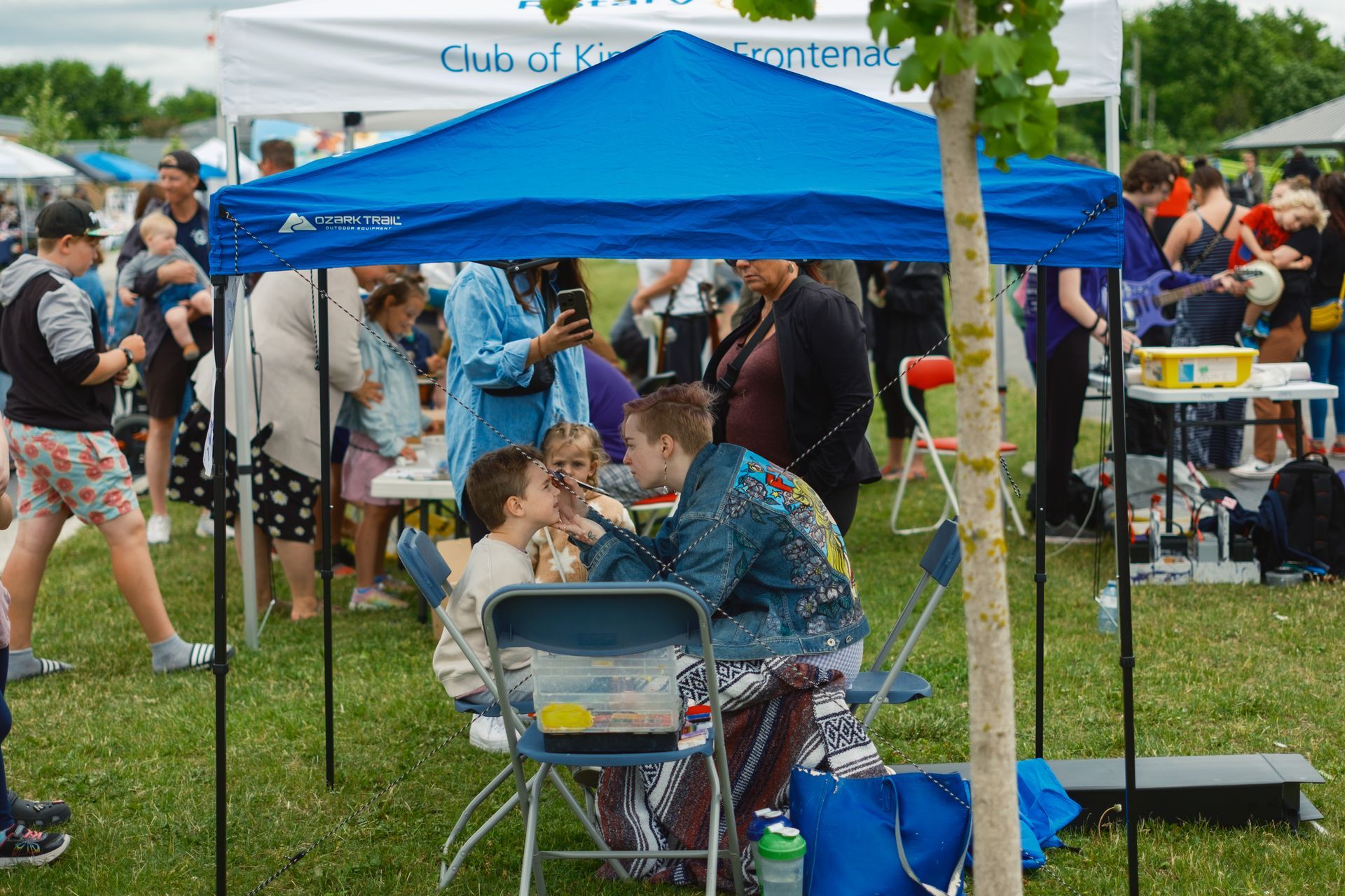 A group of people are sitting under a blue tent in a park.
