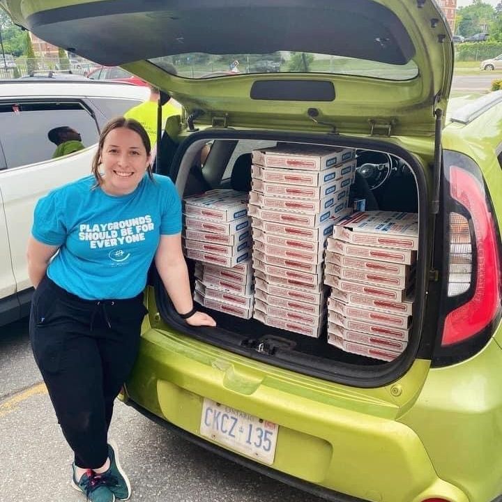 A woman in a blue shirt stands next to a green car filled with pizza boxes