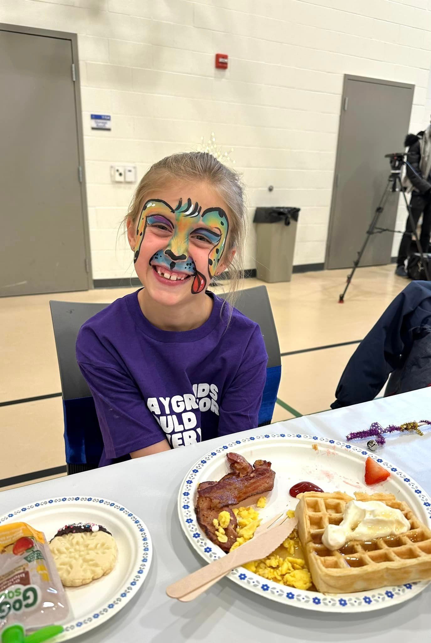 A young girl with face paint is sitting at a table with plates of food.