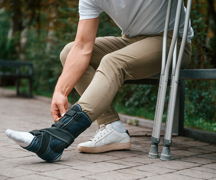 A man is sitting on a bench with crutches and tying his ankle brace.