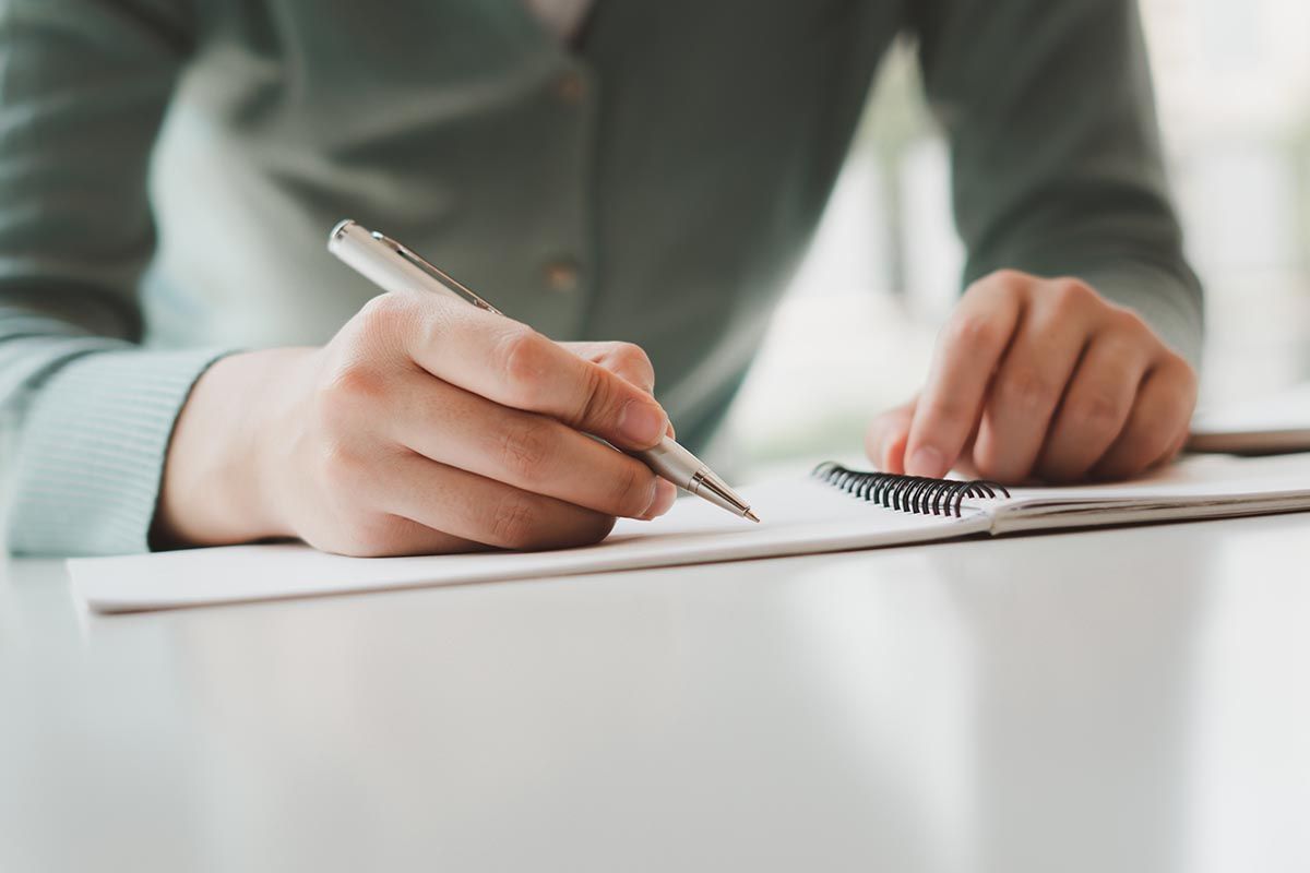 A person in a light green sweater writes on a notebook with a silver pen at a white desk.