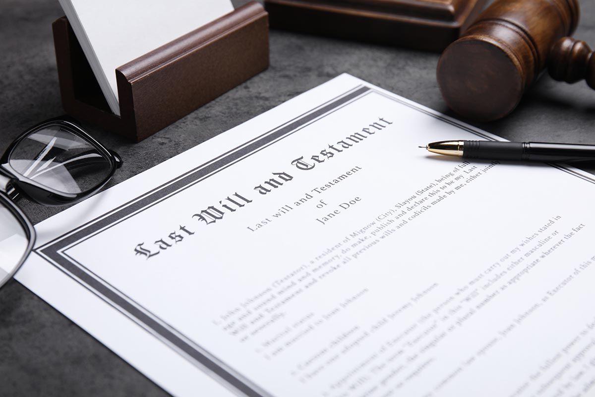 A document titled Last Will and Testament sits on a dark desk with a wooden gavel, eyeglasses, and a pen nearby.