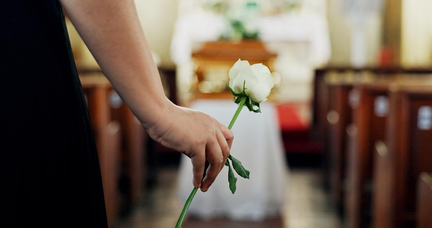 A hand holding a white rose in the foreground, with a blurred casket in a chapel setting in the background.