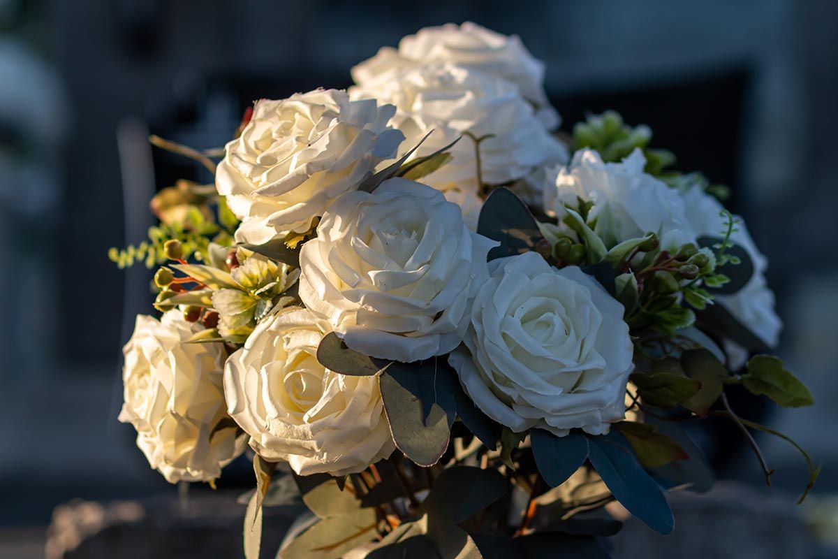 A bouquet of white artificial roses with green foliage, illuminated by warm sunlight against a dark background.