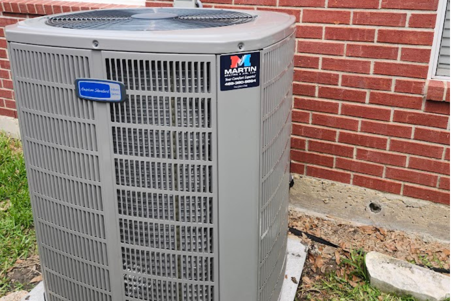 A gray air conditioner is sitting in front of a brick building.