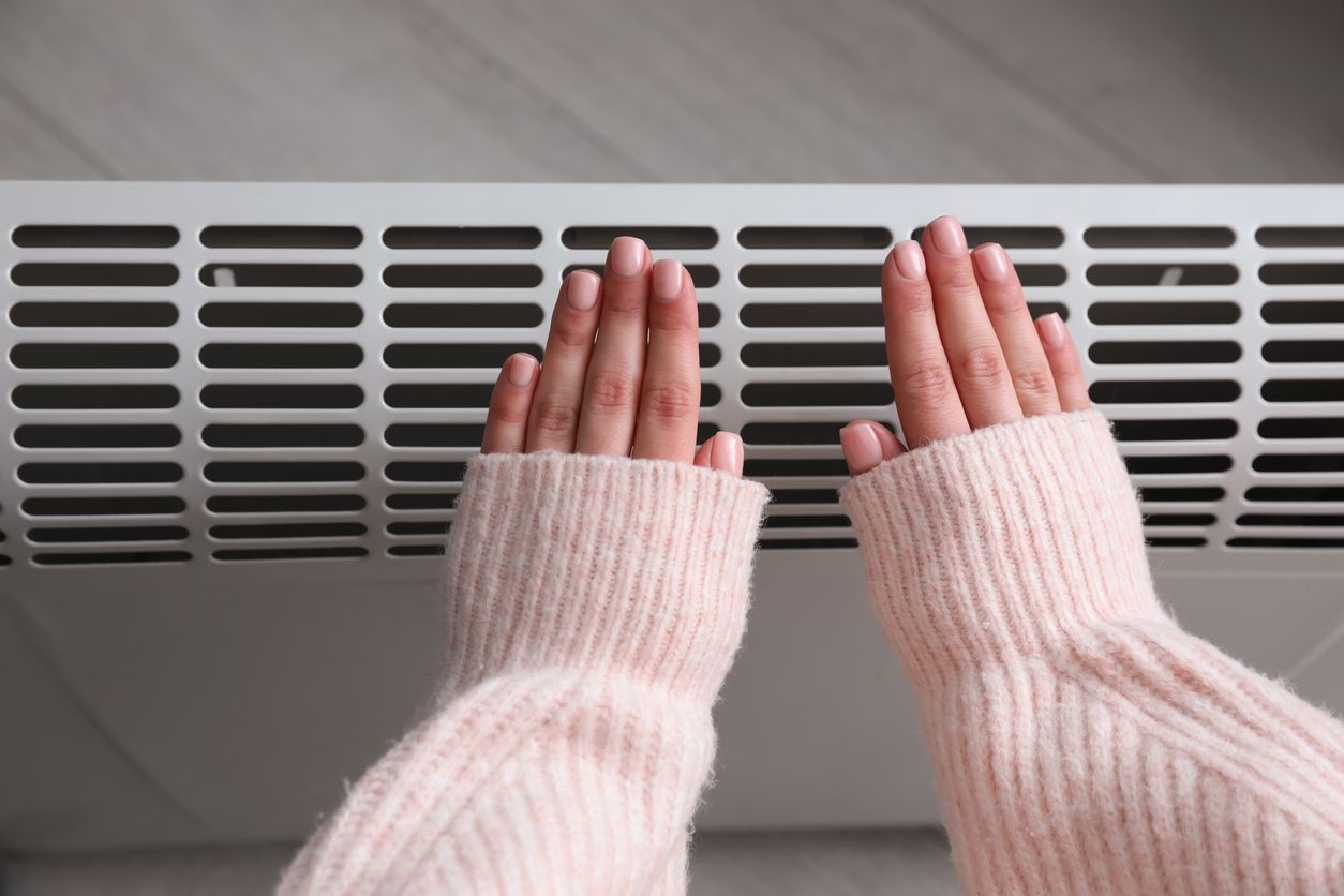 A woman in a pink sweater is warming her hands on a radiator.