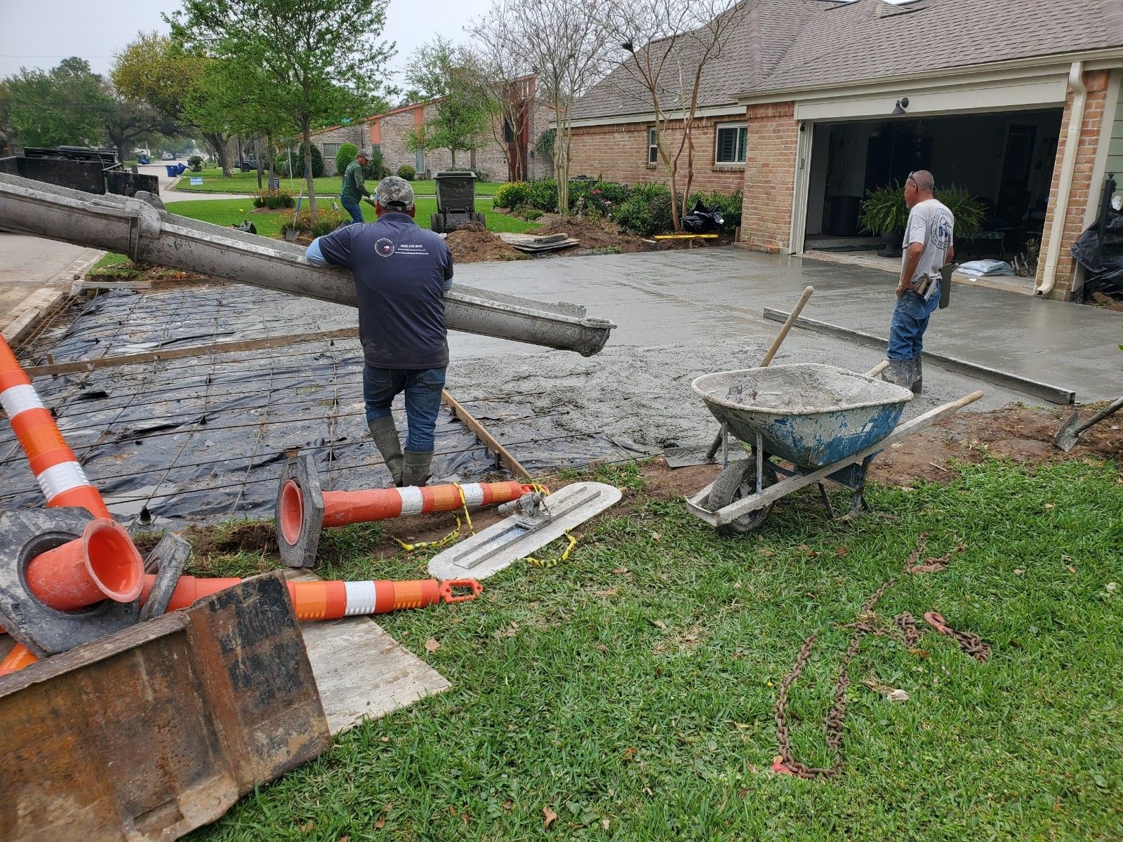 Construction workers pouring concrete driveway. Grey cement, orange cones, green grass, residential area.