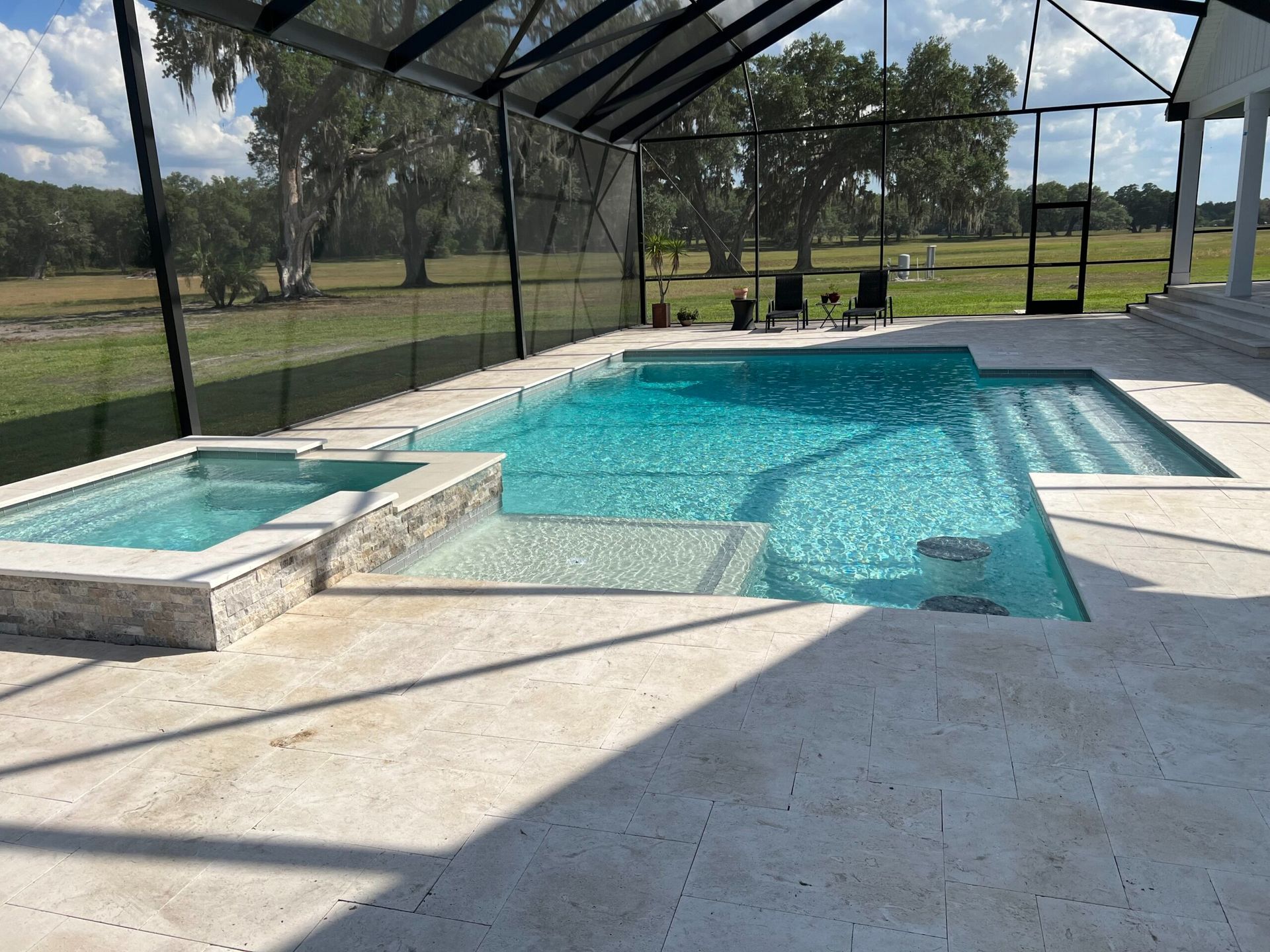 A rectangular swimming pool with a jacuzzi and steps, surrounded by light-colored stone. A screen enclosure and green field are visible in the background.