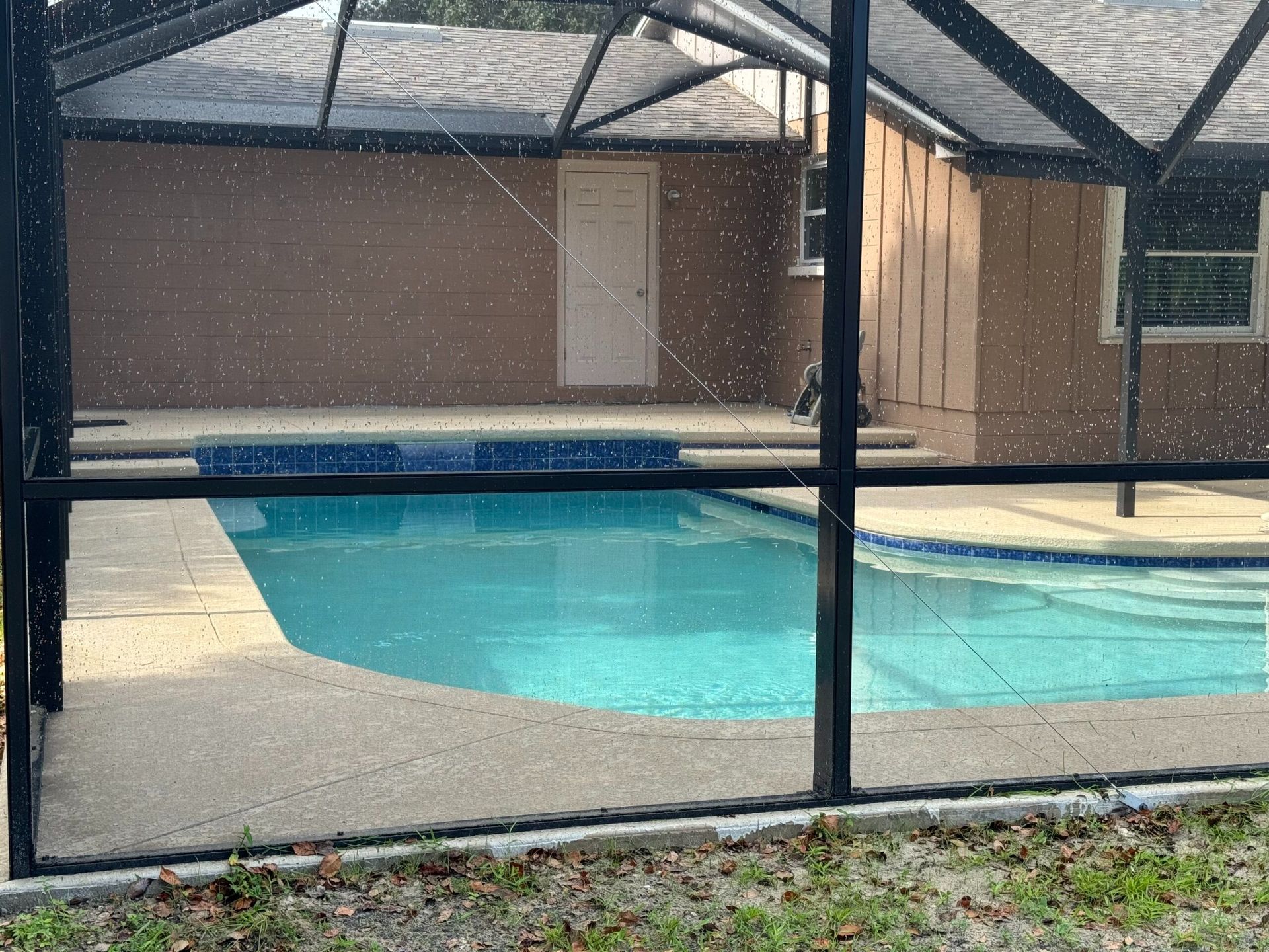 Swimming pool enclosed by a screened patio. Turquoise water, concrete deck, and a brown building in the background.