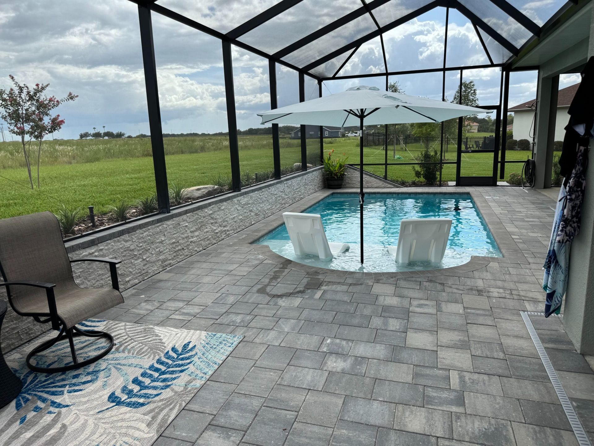 Screened-in patio with a small pool, a sun umbrella, and two in-pool chairs. Gray pavers, stone wall, and a chair on the patio.