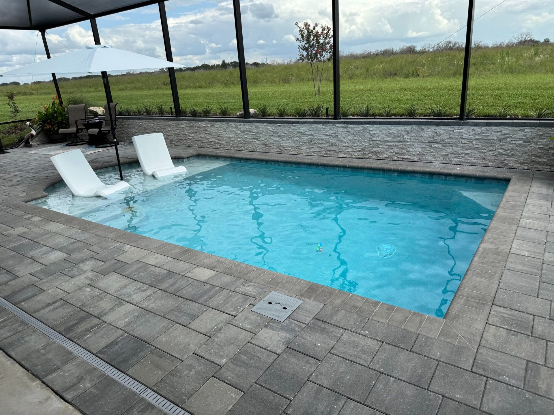 A small, rectangular pool with two white lounge chairs sits on a patio. Green field visible beyond a screen enclosure.
