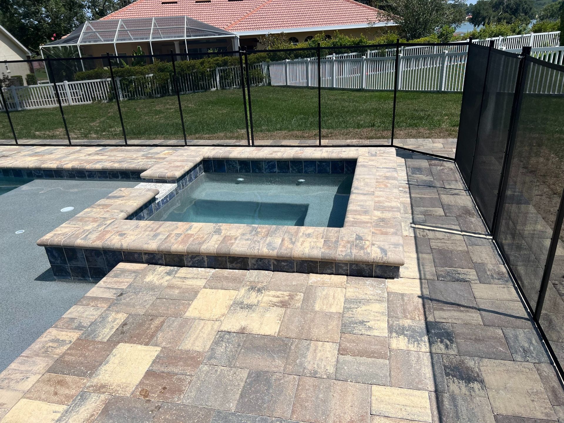 A backyard with a hot tub next to a pool, surrounded by paving stones and a black fence.