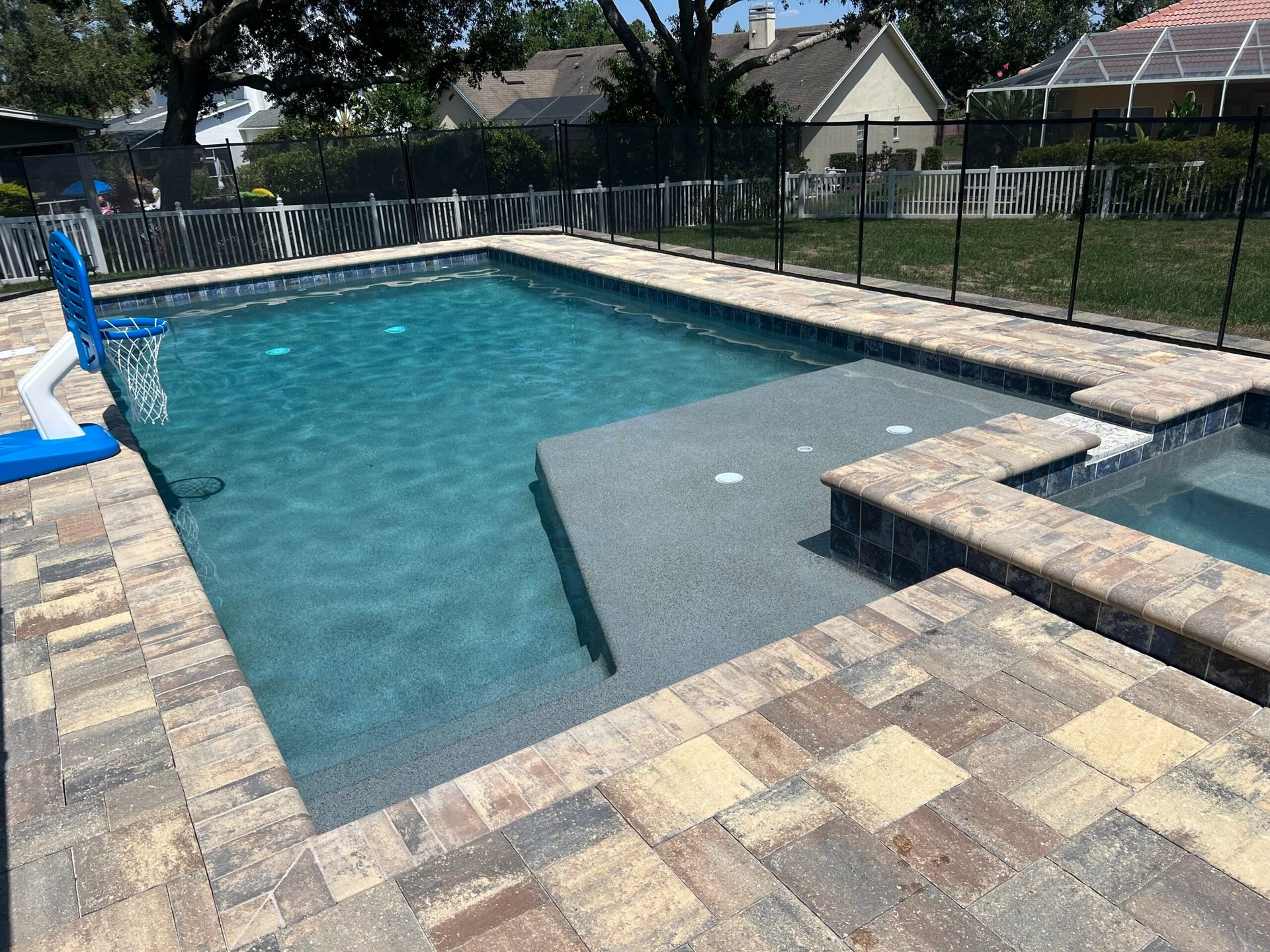 A rectangular swimming pool with a jacuzzi. The pool has blue water, surrounded by a paver patio and a black fence.