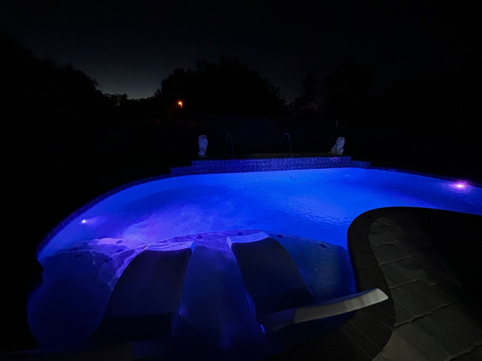 Nighttime view of a pool illuminated with blue lights. Two statues are visible on the pool's edge.