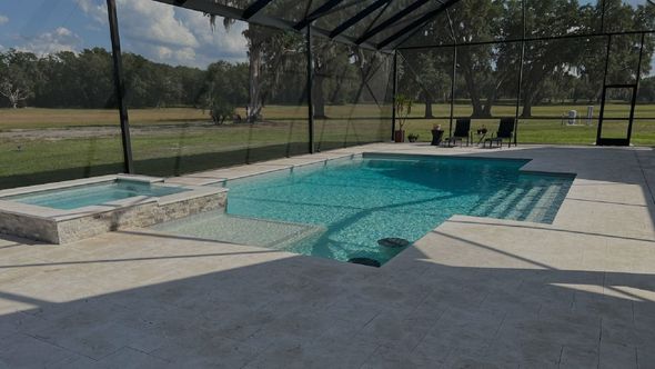 A screened-in outdoor pool and hot tub with lounge chairs on the deck, surrounded by grass and trees.