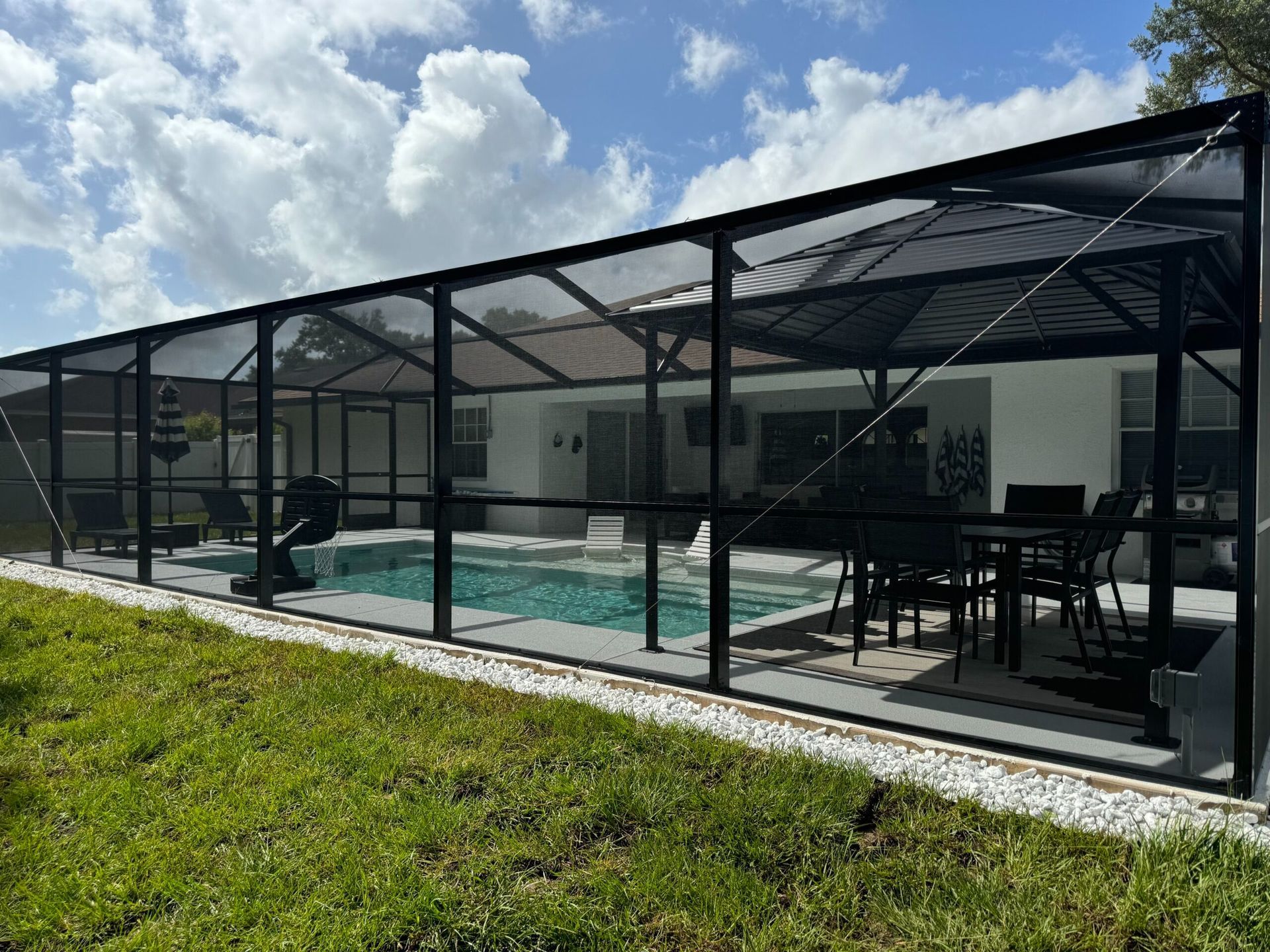 A screened-in pool enclosure with black framing. The enclosure surrounds a pool with seating and a table inside, set against a white house with a green lawn.
