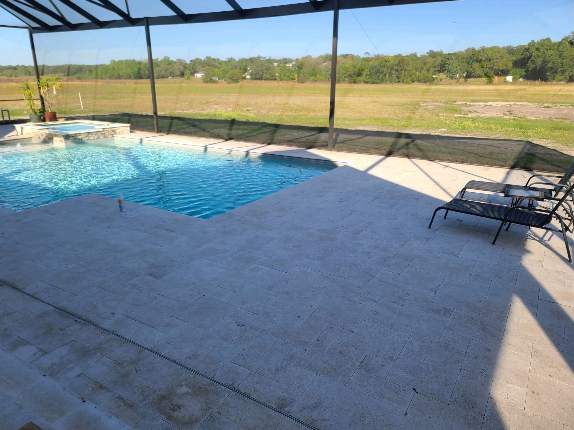 Poolside view with a blue pool, concrete patio, and two lounge chairs under a screened enclosure, overlooking a grassy field and trees.
