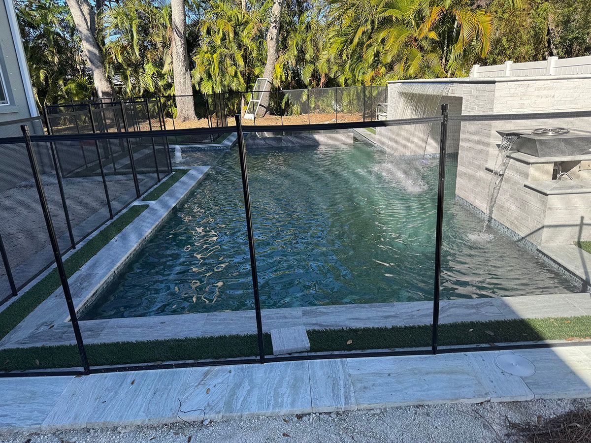 A black mesh pool fence surrounds a backyard pool with a waterfall feature. The water is a light blue-green.