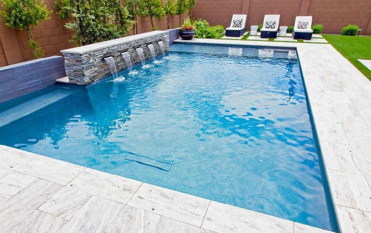 A rectangular swimming pool with stone waterfall feature and lounge chairs on a patio. The water is bright blue.