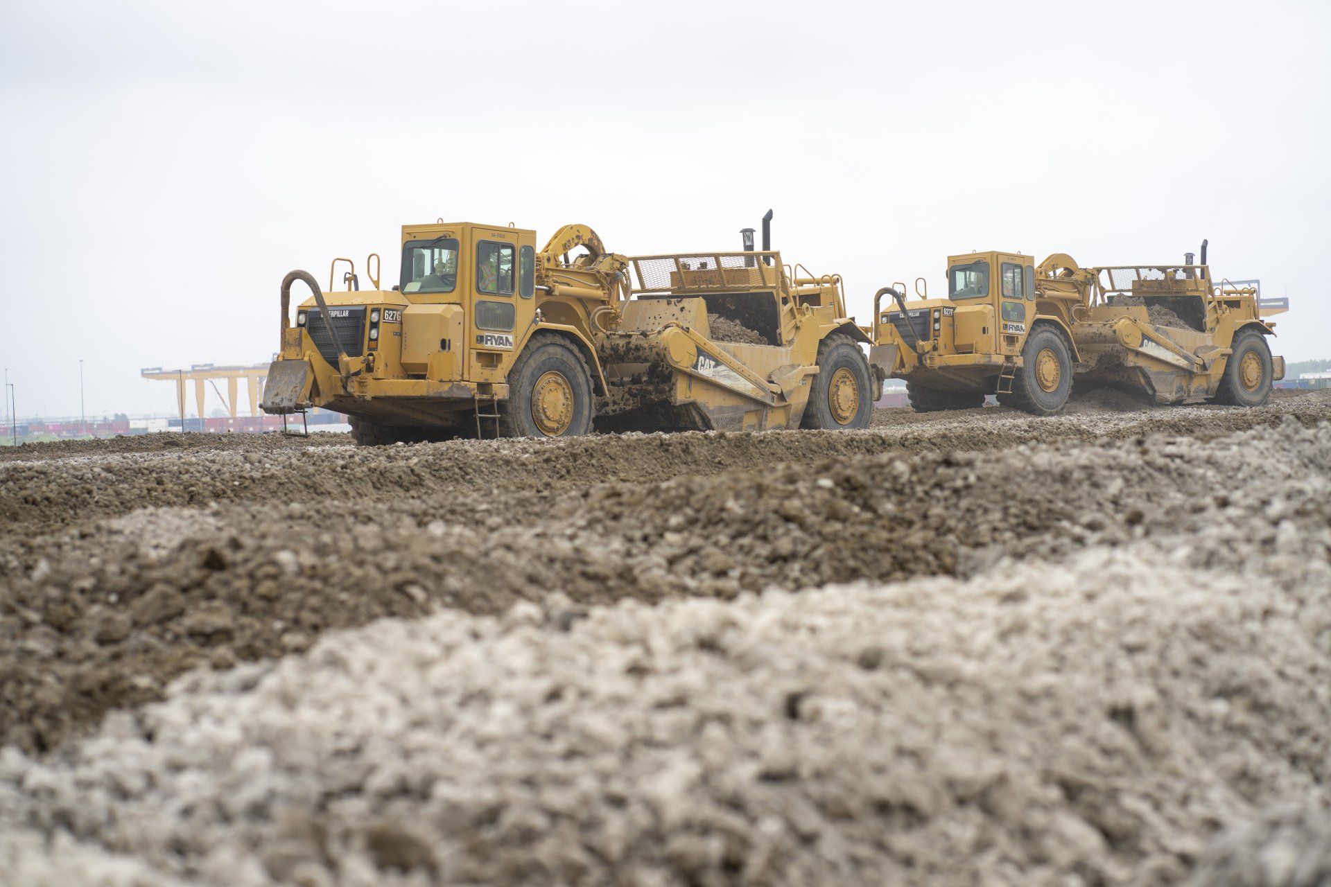 A row of construction vehicles are driving down a dirt road.