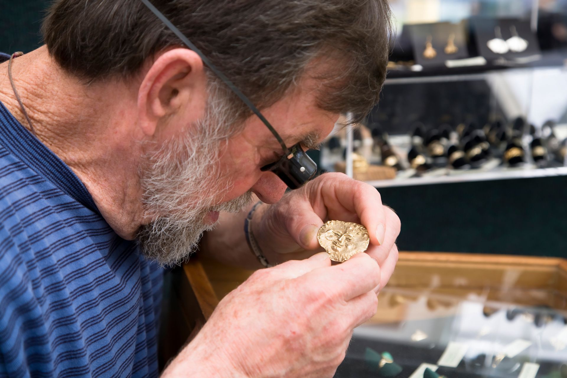 Jeweler inspecting a gold nugget with a loupe in a store display of rings and jewelry.