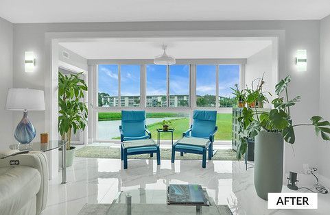 Bright living room with blue chairs facing large windows overlooking a lake and buildings; plants and light fixtures are present.