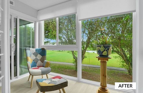 Sunroom with large windows, colorful patterned chair, ottoman, and plant on a wooden pedestal overlooking a green yard.