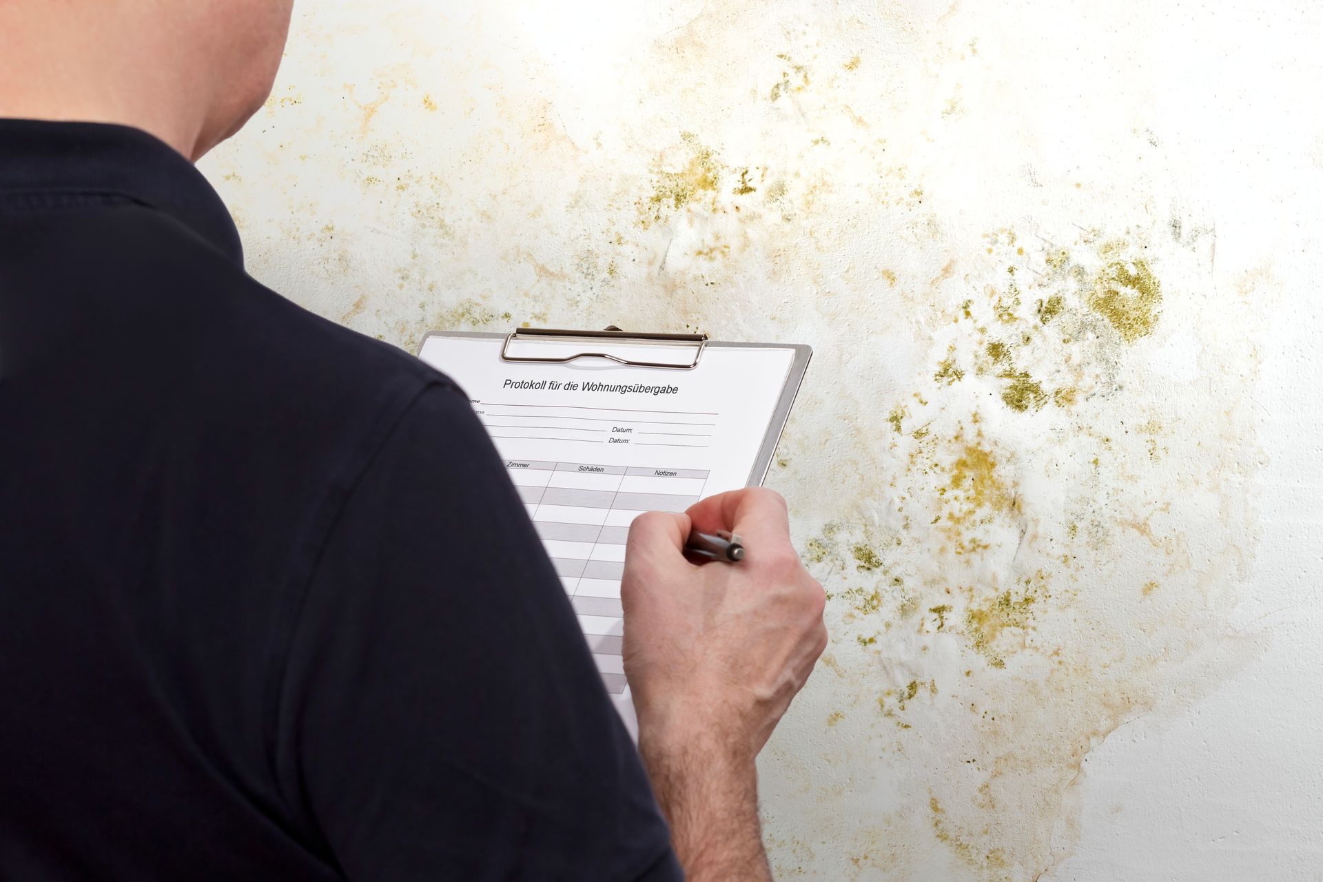 A man is writing on a clipboard in front of a mouldy wall.