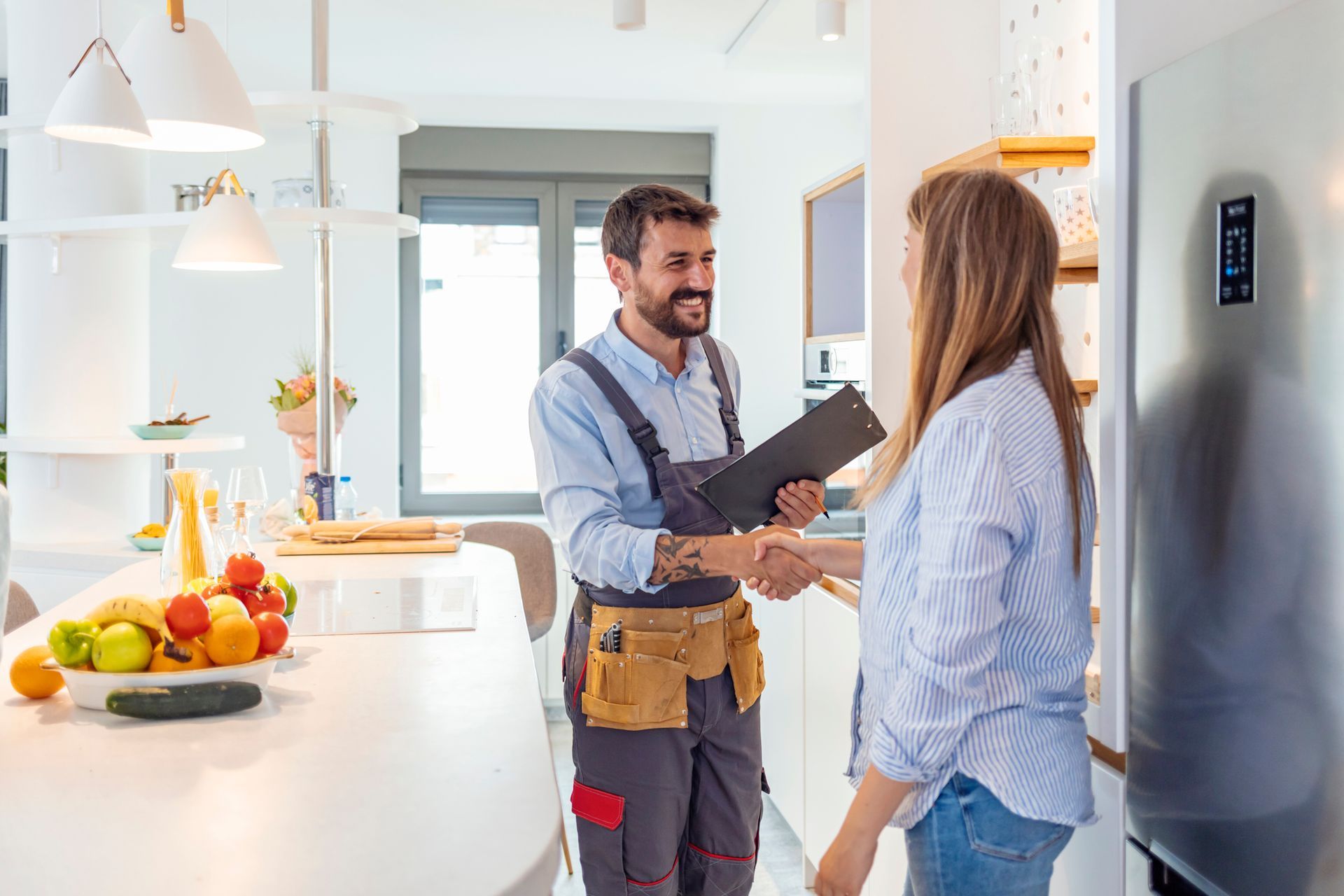 A woman shakes hands with a residential plumbing contractor, holding a clipboard, in the kitchen.