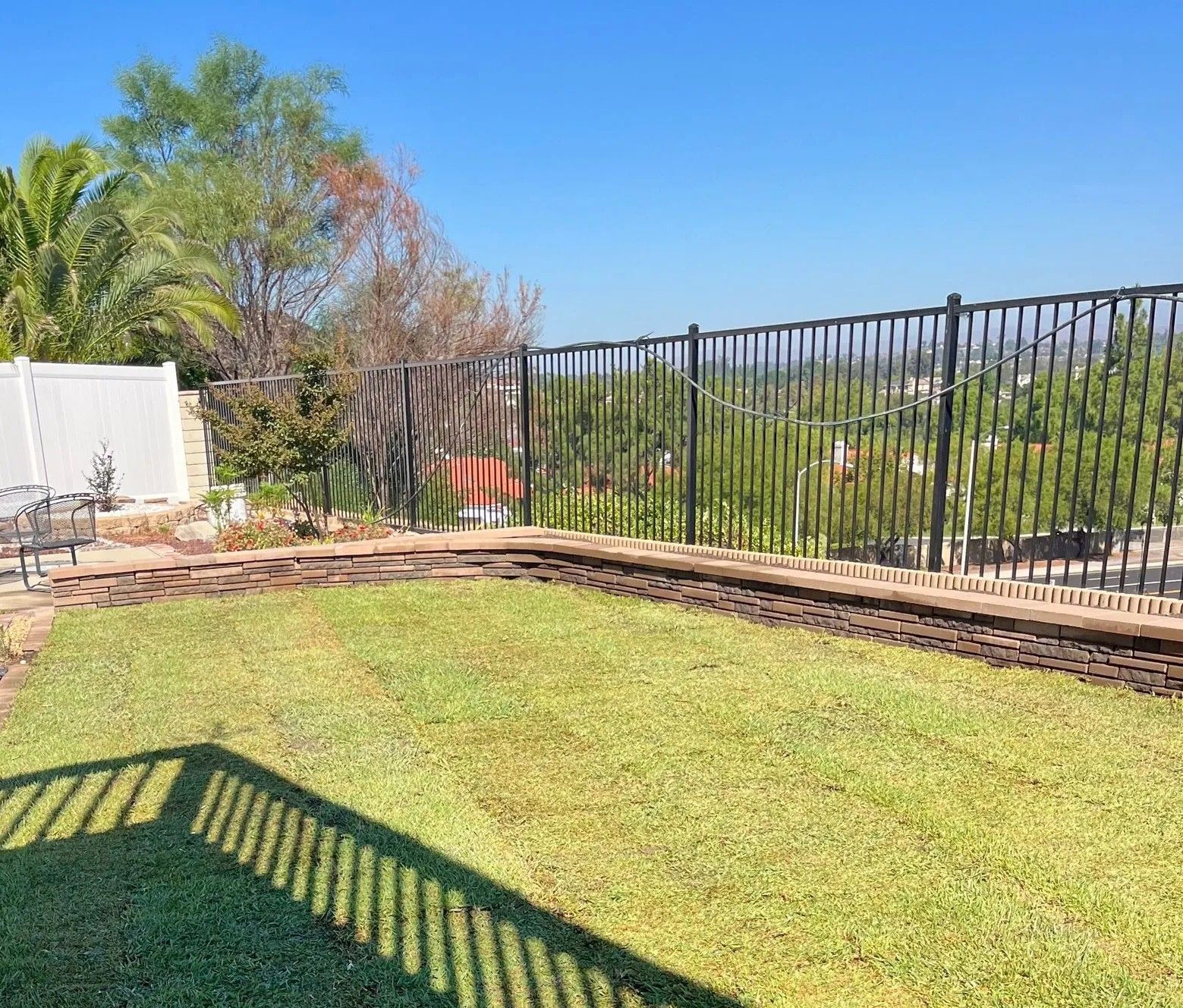 Green lawn in a backyard, bordered by a retaining wall. Black metal fence overlooks a distant view. Blue sky.