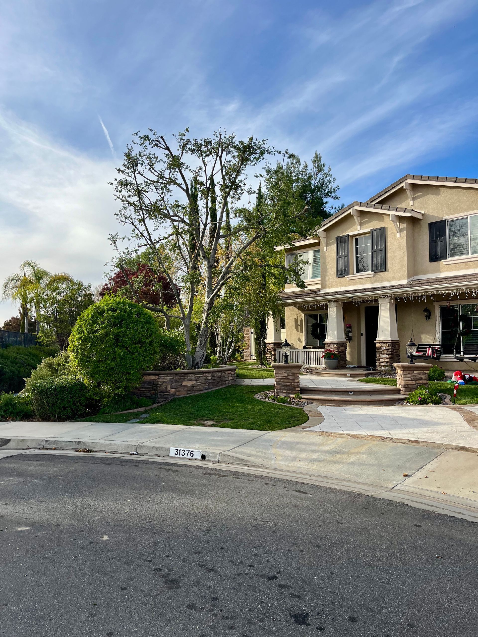 Suburban house with stone facade, front porch, trees, and a curved driveway under a bright sky