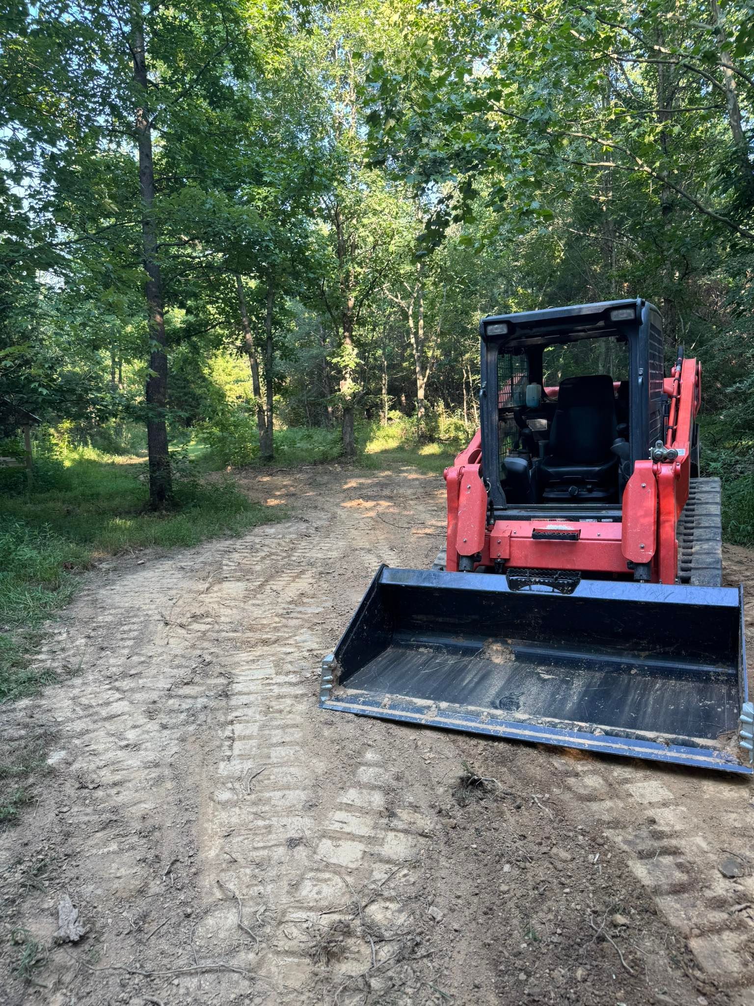 Orange and black skid steer on a dirt path in a wooded area. The path is muddy, and trees surround the scene.