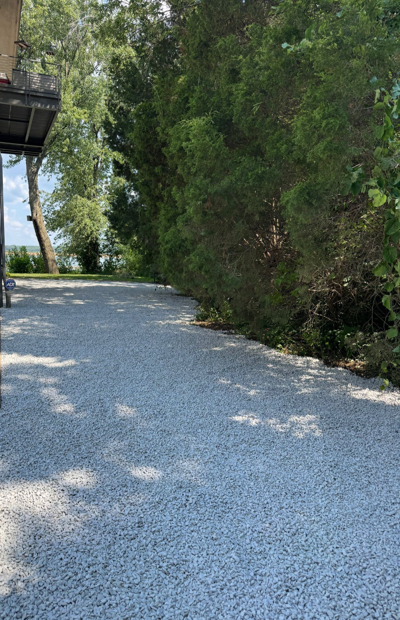 Gravel driveway next to a building and dense green bushes, leading towards a glimpse of water and trees.