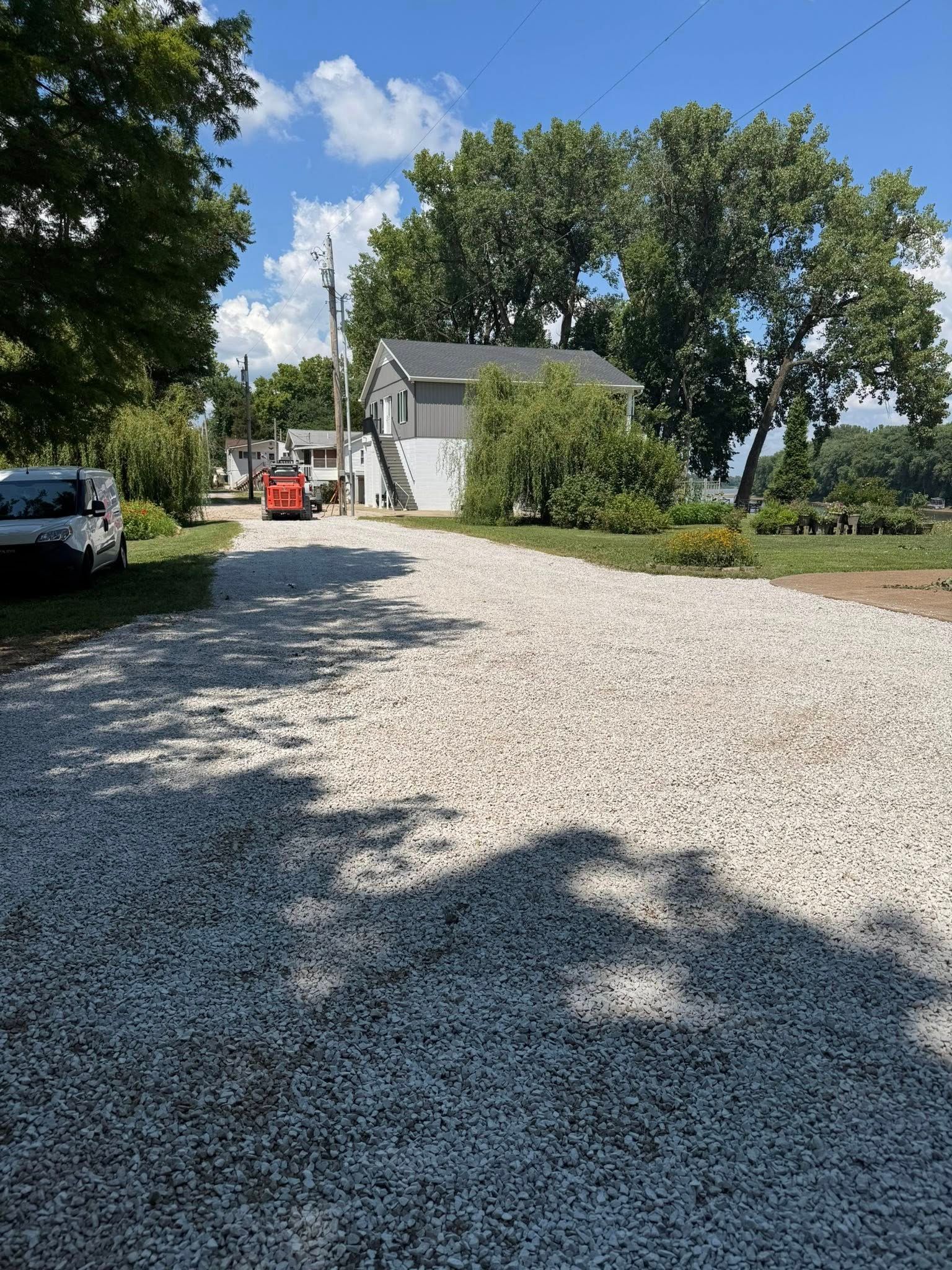 Gravel driveway leads to a gray house under a blue sky. A red car is parked halfway up.