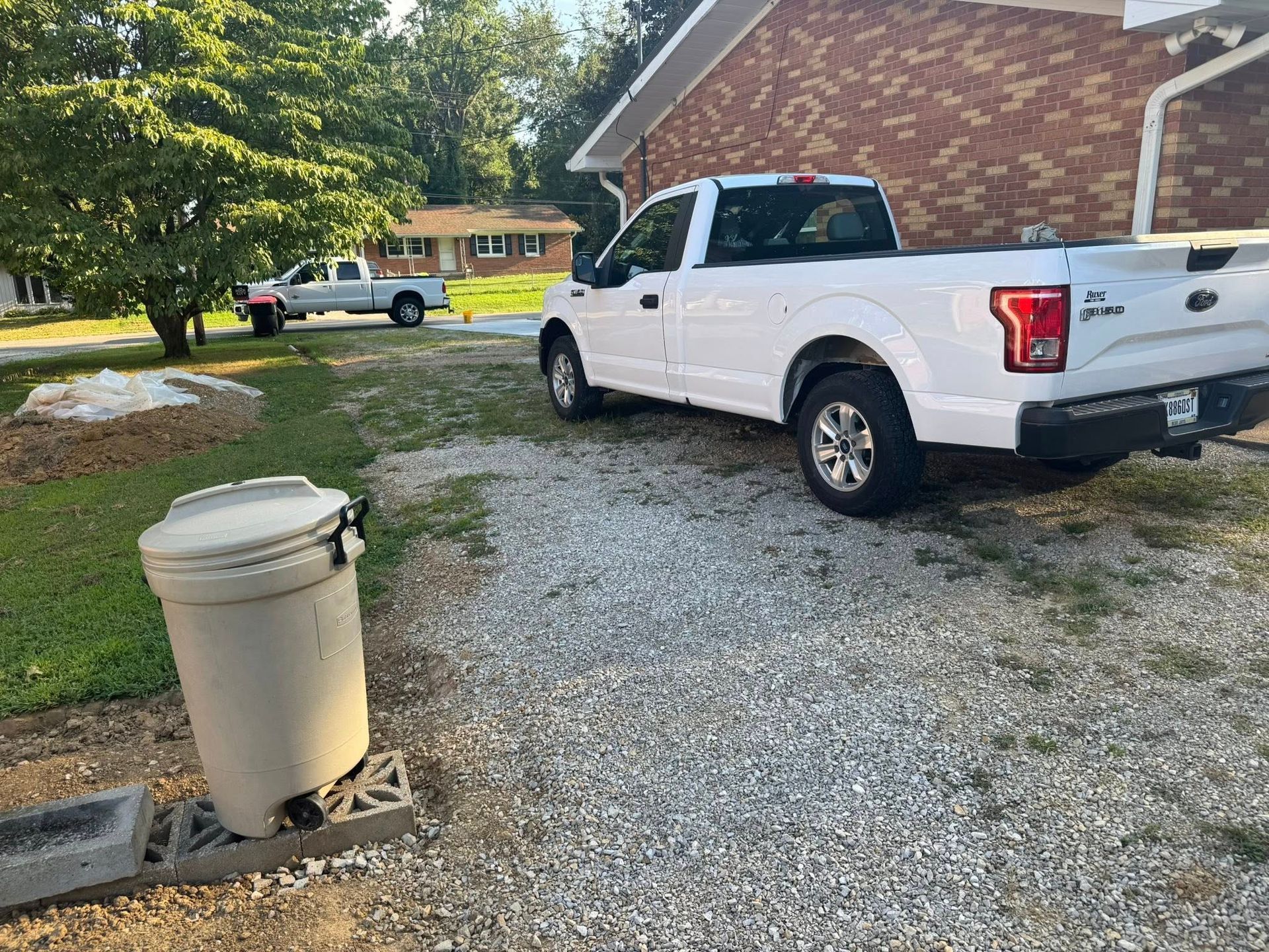 White pickup truck parked in a gravel driveway, trash can in foreground, another truck in the distance.