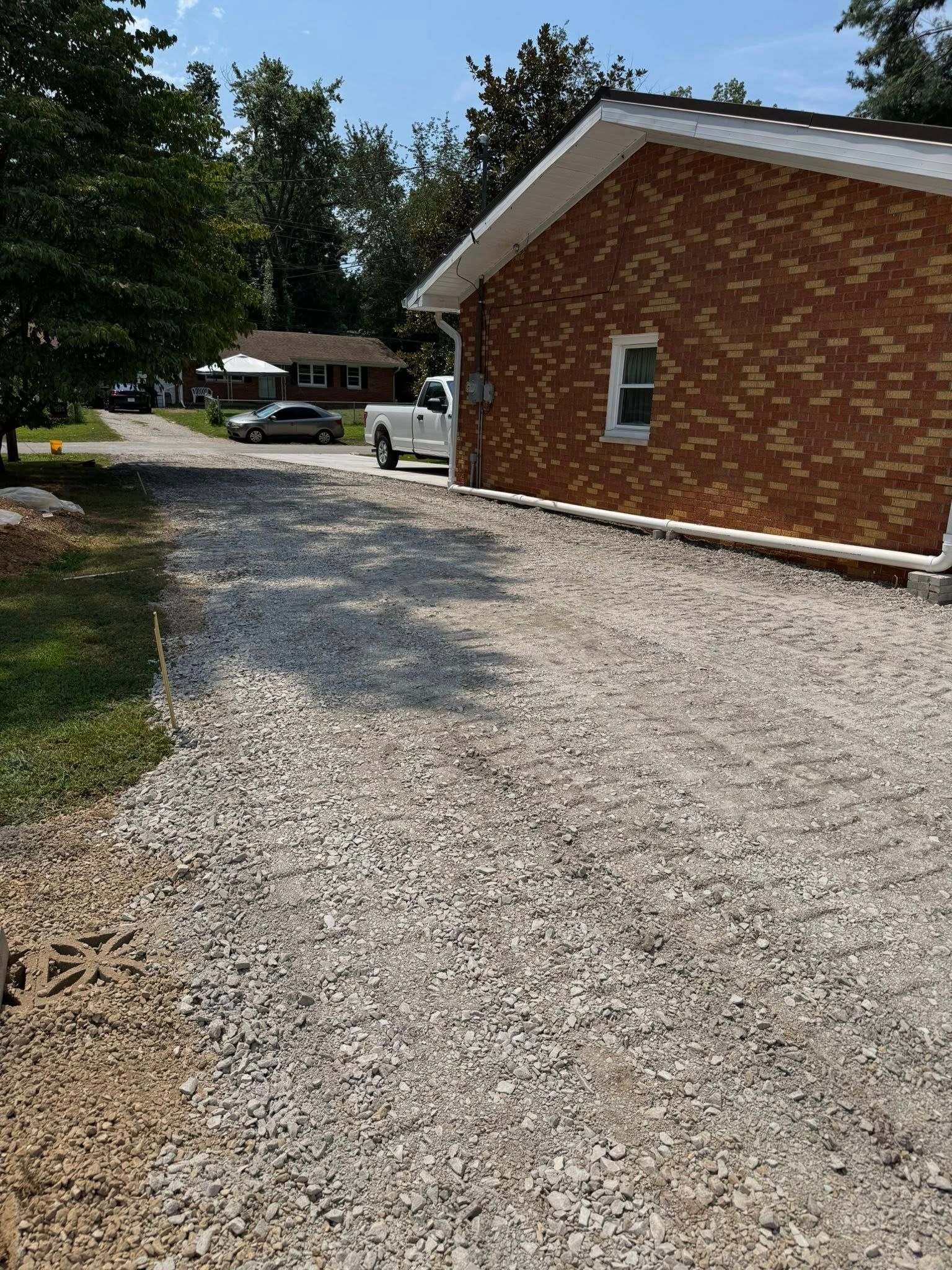 Gravel driveway next to a brick building. Cars and a house are in the background. Sunny day.